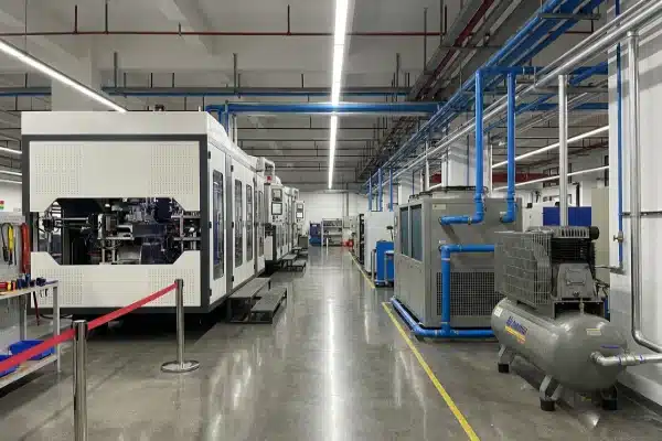 A detailed industrial perspective of a plastics factory aisle featuring a water chiller and air compressor connected with color-coded piping to support an extrusion blow molding production line.