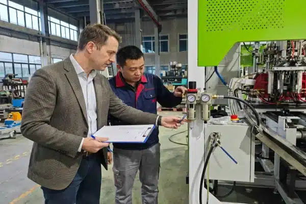 A Western business owner and a Chinese project manager conducting a Factory Acceptance Test (FAT) by inspecting the FRL assembly and automatic drain on an all-electric blow molding machine.